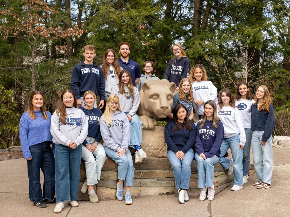 Seventeen college students wear Penn State apparel while sitting or standing around the Nittany Lion Shrine.