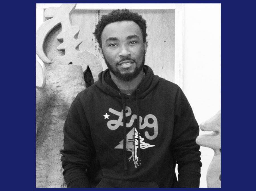 Black and white image of a young Black man standing in an art studio smiling