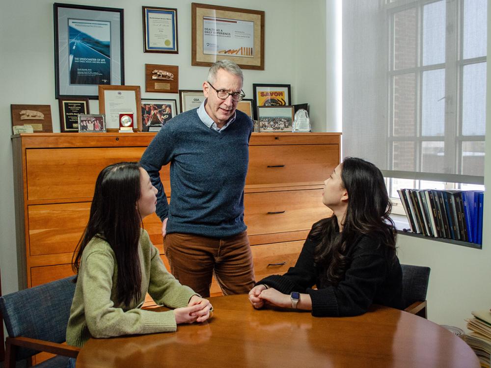 David Almeida (center) meets with two graduate students in his office to discuss research. They are seated at a round table and a bookshelf full of books and items is behind them.