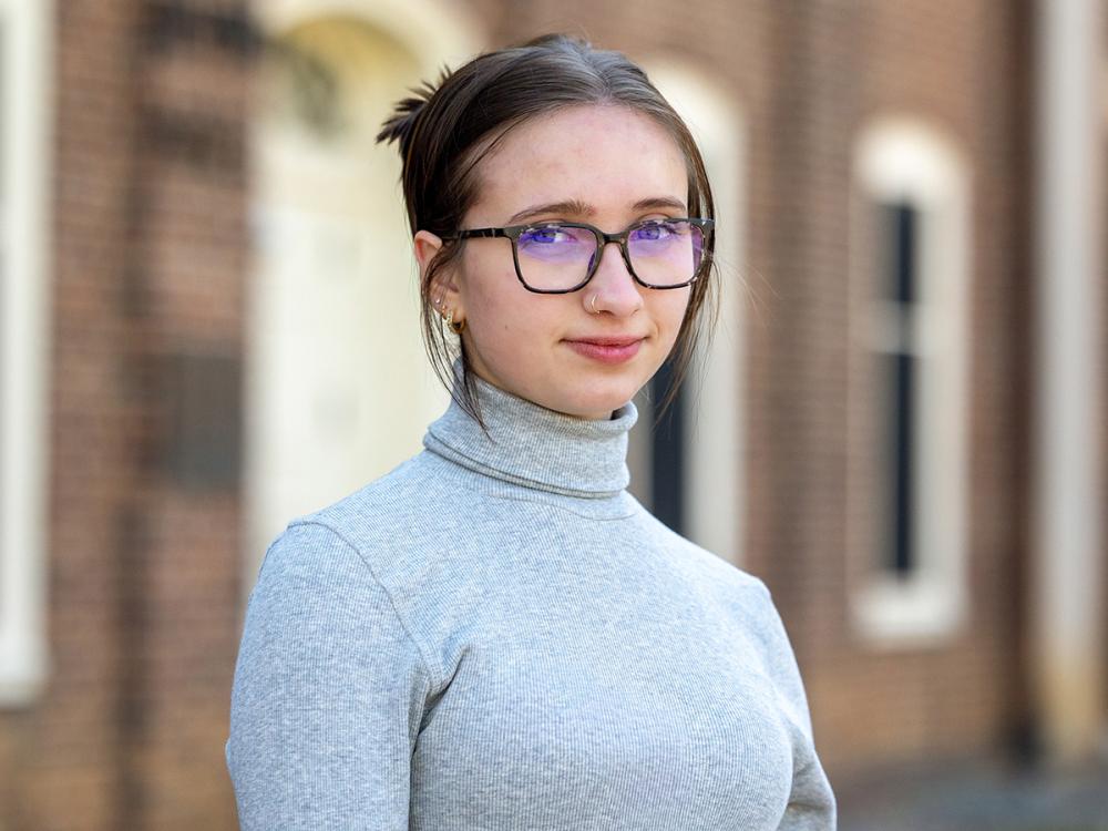 Amaya wears glasses and a gray turtleneck stands confidently outdoors, brick wall and windows in the background.