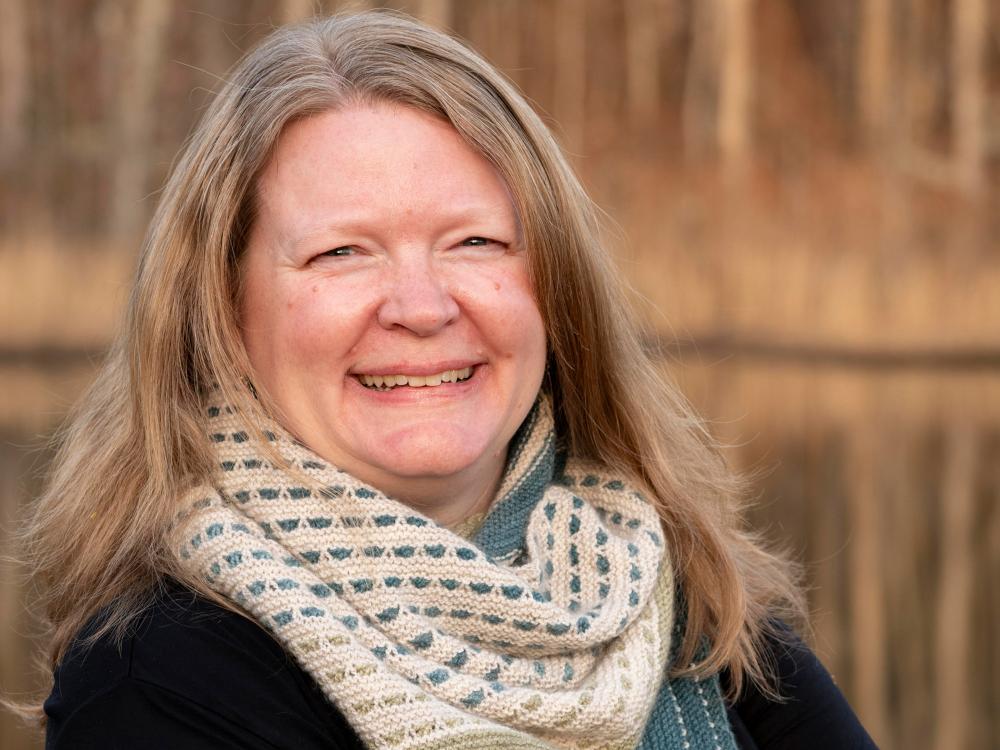 Person with shoulder‑length hair wearing a patterned scarf and dark top, standing outdoors in front of a background of trees.