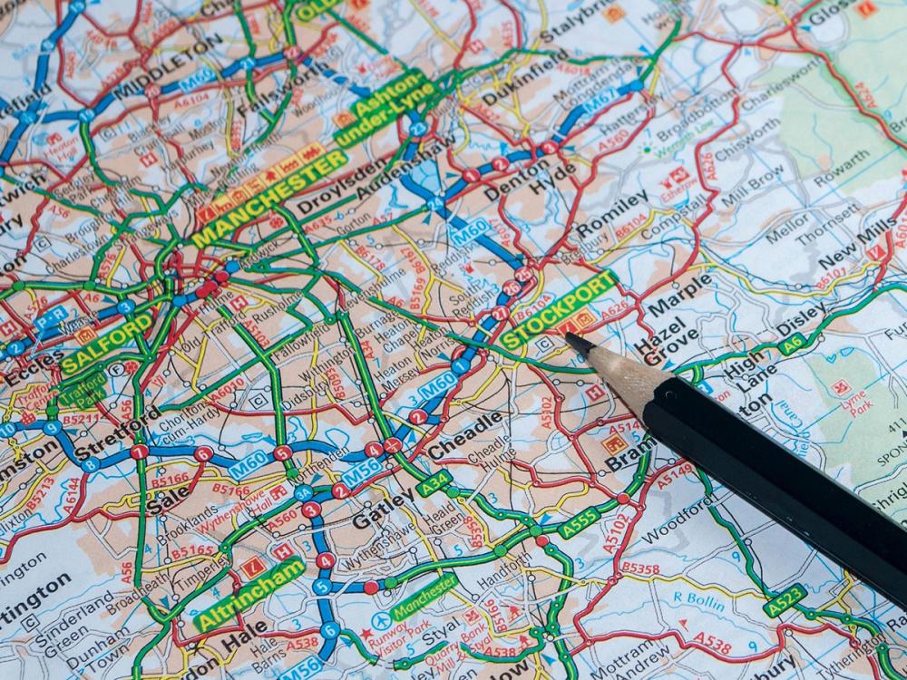 A detailed road map showing Manchester, Stockport, and surrounding areas, with roads highlighted in red and green. A pencil points to Stockport.