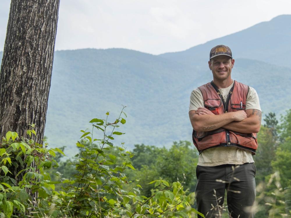 Ethan Tapper, a man in a red vest, yellow ballcap and khaki pants, stands next to a tree in the woods with mountains in the background.