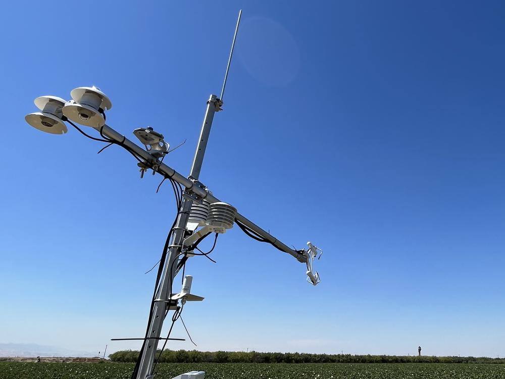 A cross-shaped piece of equipment with attached sensors next to a field of crops against the blue sky