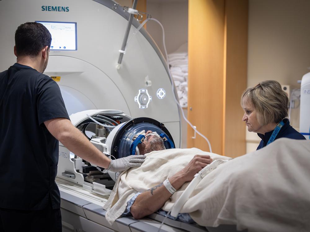 Two MRI technologists stand next to a male patient who is about to go into an MRI machine. The patient has a headgear on his head and a nasal cannula under his nose. 