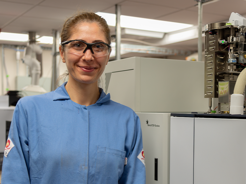 Hilal Ezgi Toraman, wearing safety glasses and a blue lab coat, is smiling while standing in a research lab.