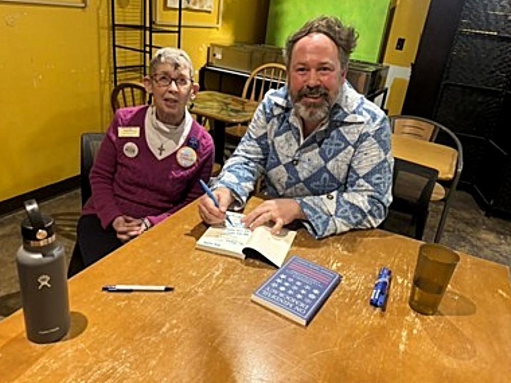Jeremy Engels, a man wearing a blue and white shirt with a gray beard and signing his book, sits next to Joan Bouchard, a woman with short gray hair and a pink turtleneck with a pink v-neck sweater.