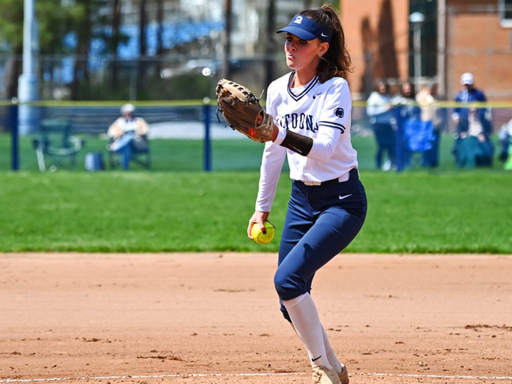 Softball player Josselyn Nau fields the ball on a sunny day, wearing a white jersey and visor, with a grassy outfield and spectators in the background.