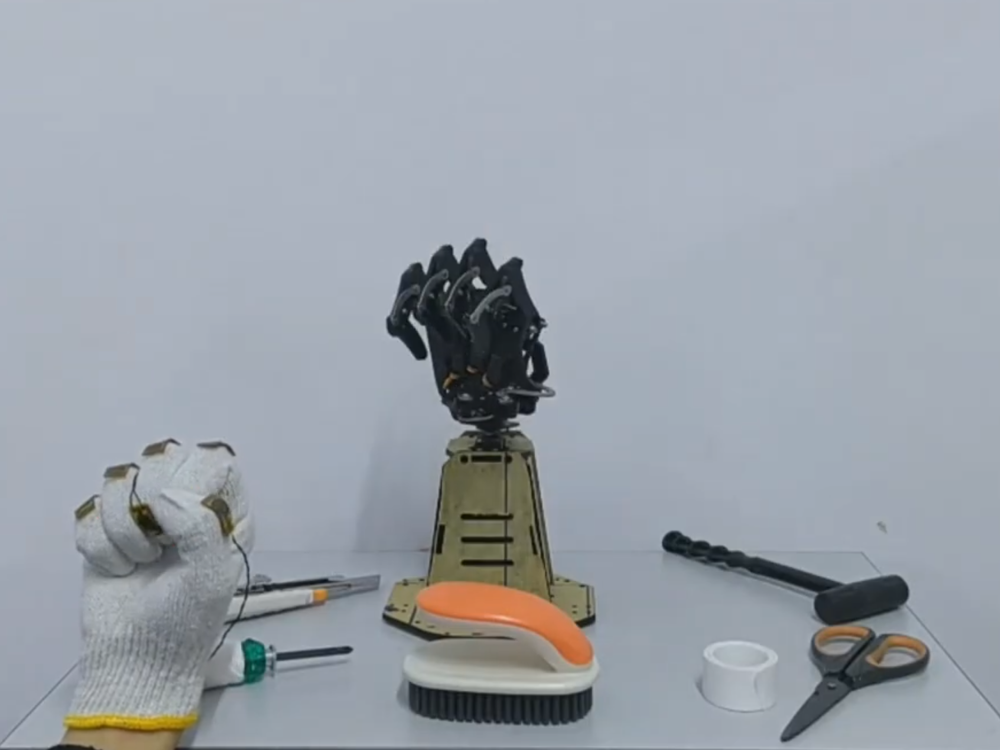 A video demonstration showing a hand in the corner controlling a robotic prosthetic hand. The robot grips and moves some household objects, including a brush and a knife. 