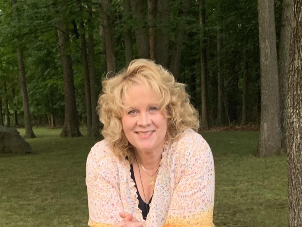 Woman with curly blonde hair leaning on a large rock in a forested park area during daytime.