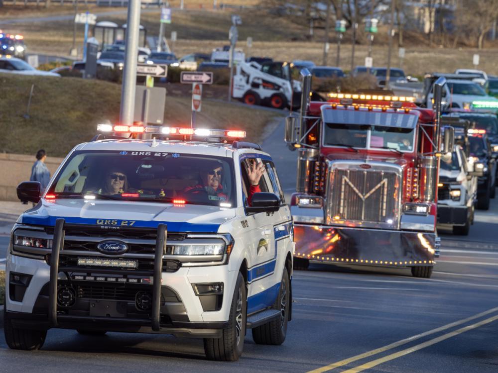 A line of vehicles with flashing lights drives toward the camera. They include emergency vehicles, tow trucks and others.