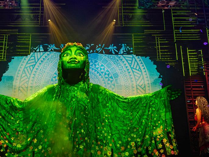 A theatre stage set with a large green goddess being looked at by a woman with brown curly hair