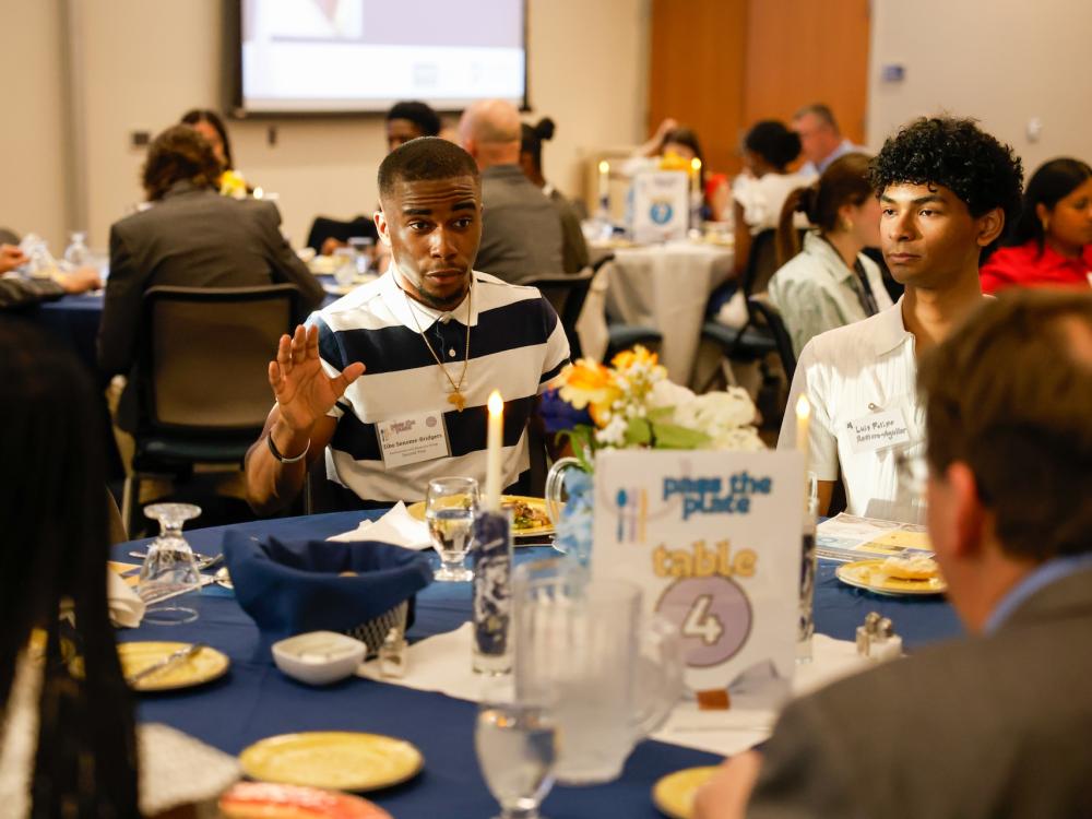 student talking at a table with blue tablecloth and "pass the plate table 4" sign at the table
