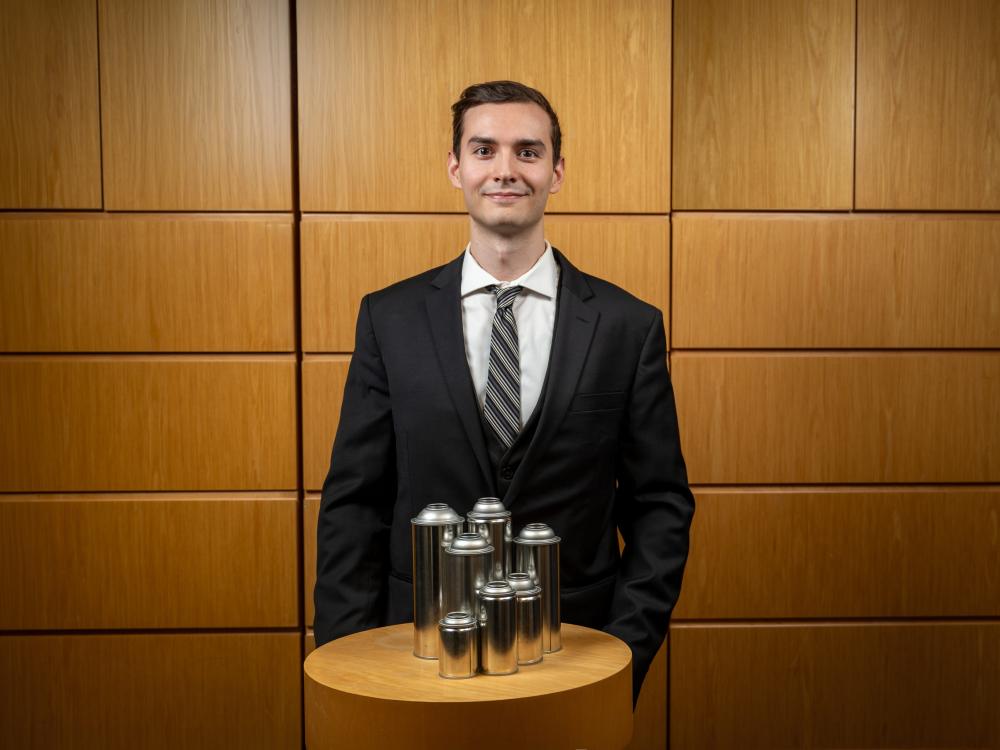 A man in a suit and tie stands behind a collection of seven steel aerosol cans of various sizes.