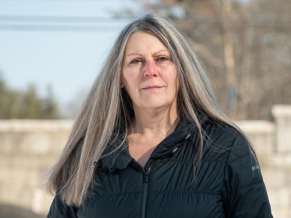 Headshot of Laurene Allen, a woman with long gray hair wearing a black down jacket and standing outside with trees in the background.