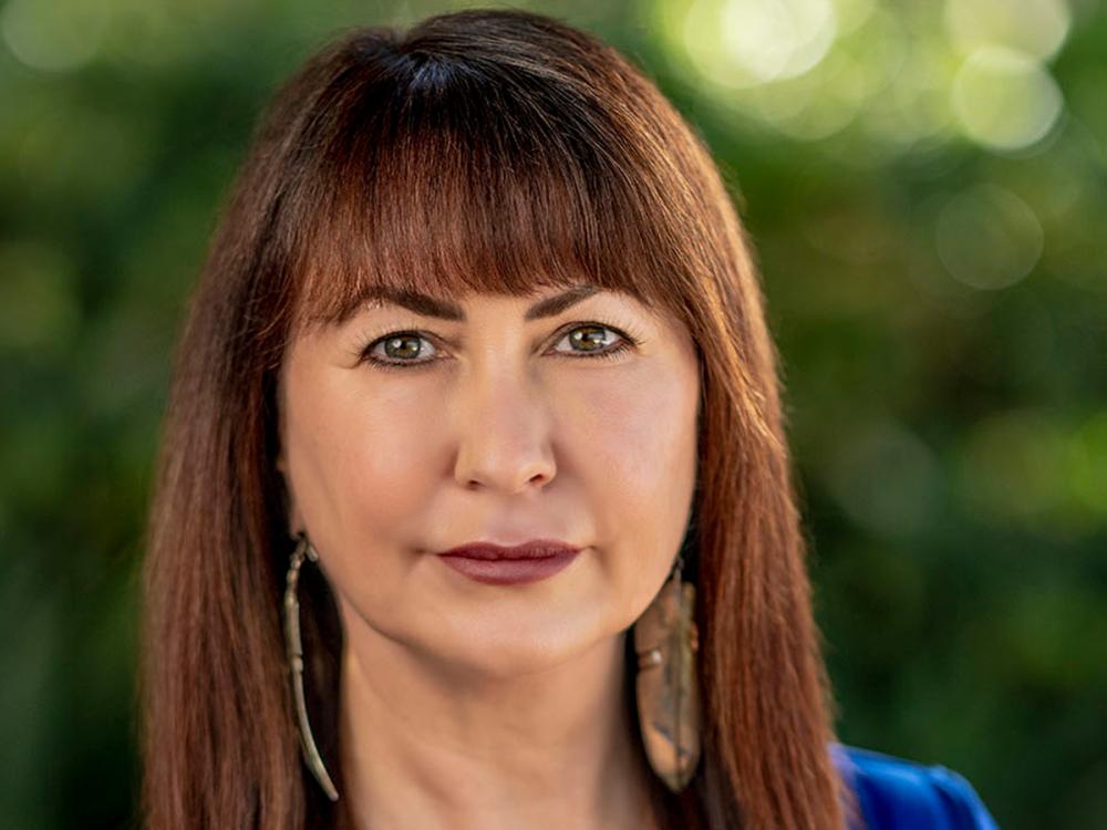 Shannon speed has long brown hair and bangs wears a blue top and large earrings, standing against a softly blurred green background.
