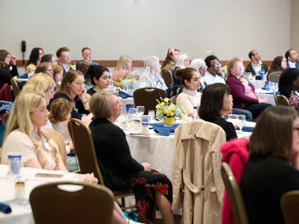Diverse groups of people sit at round tables with floral centerpieces during a formal indoor event.