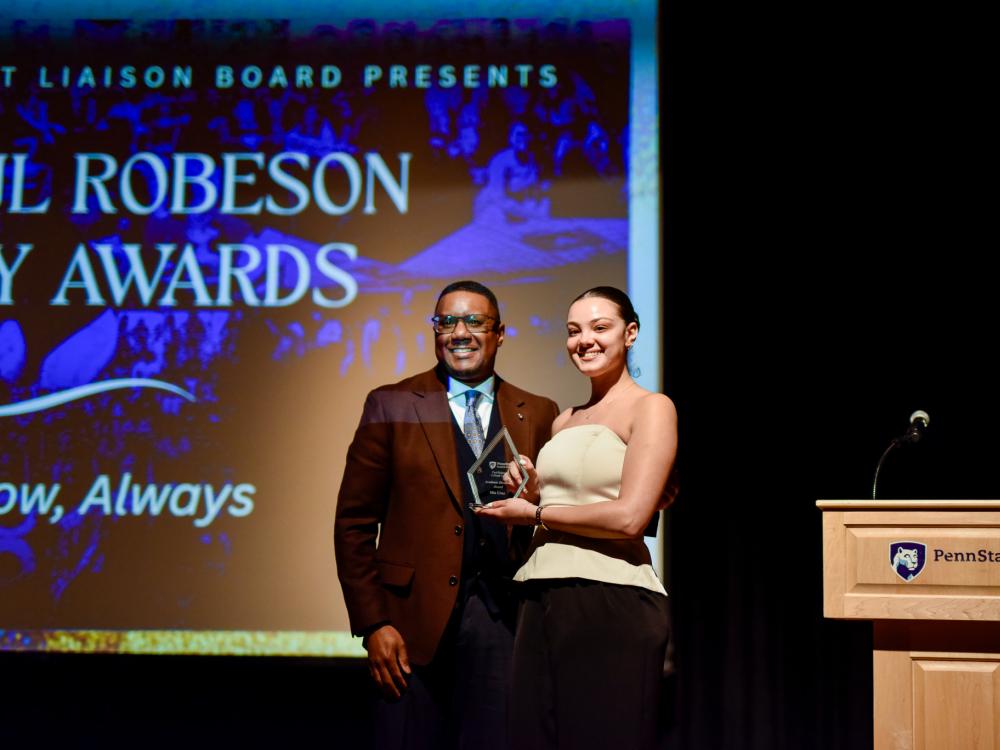 A picture of Clarence Lang, dean of the College of Liberal Arts, and Mia Cruz, the winner of the Academic Excellence Award, on the stage at Heritage Hall in the HUB-Robeson Center. Mia is receiving her award.