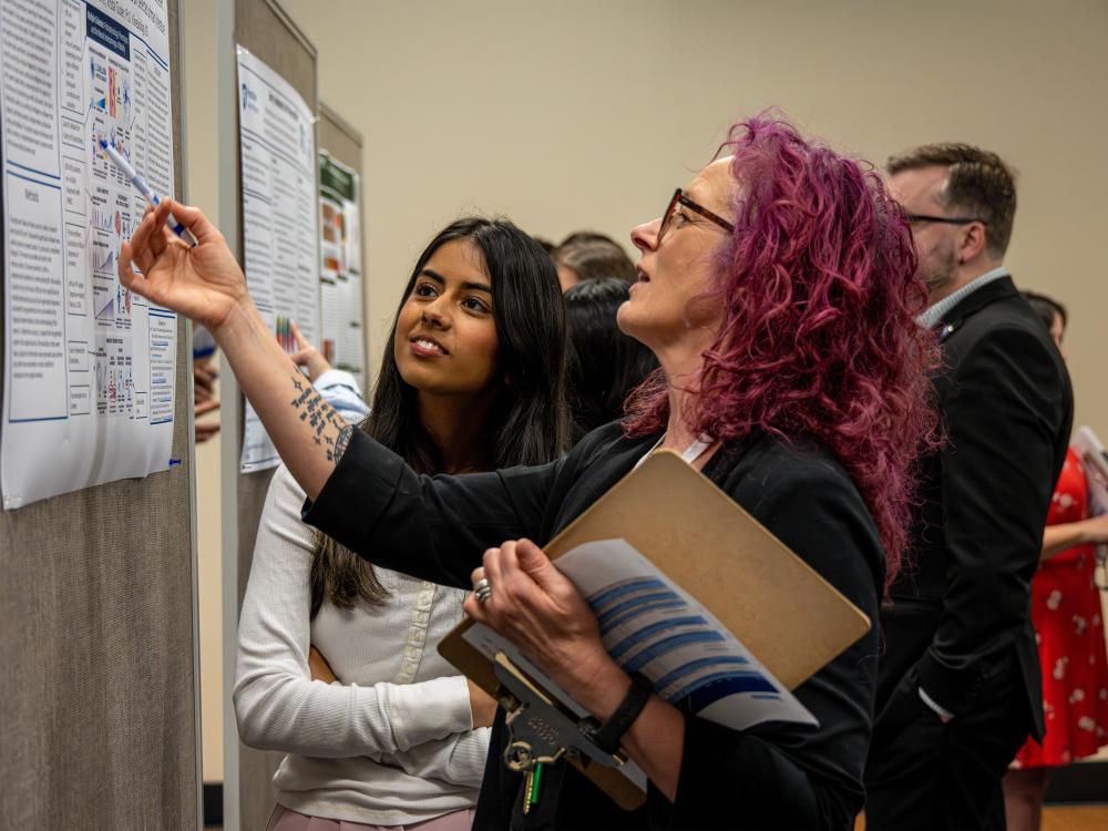 Two people stand in front of a research poster displayed on a bulletin board in an indoor academic setting. One person holds a clipboard and gestures toward the poster while the other stands nearby, with additional posters and attendees visible in the background.