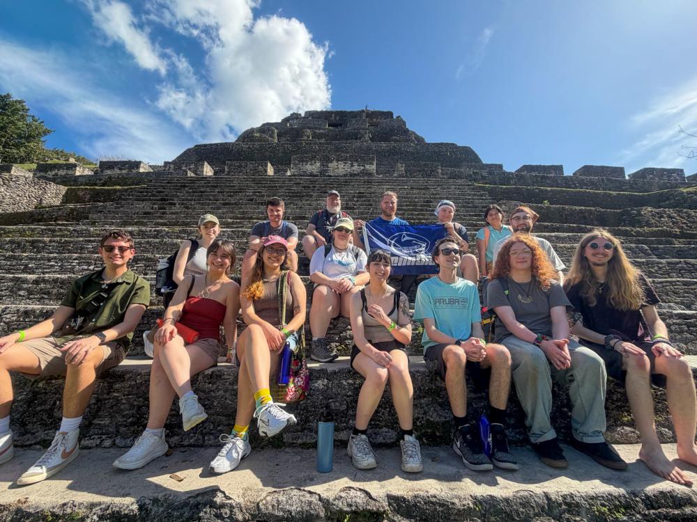 A group of fifteen people sit together on the stone steps of an ancient pyramid at an archaeological site. One person in the back row holds a blue-and-white Penn State flag. The sky is bright and clear, and the group is dressed in casual warm weather clothing.