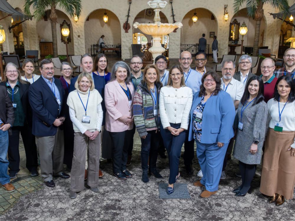 A group of approximately 23 people in business casual attire pose for a group photo in a hotel lobby. A fountain is in the near background.