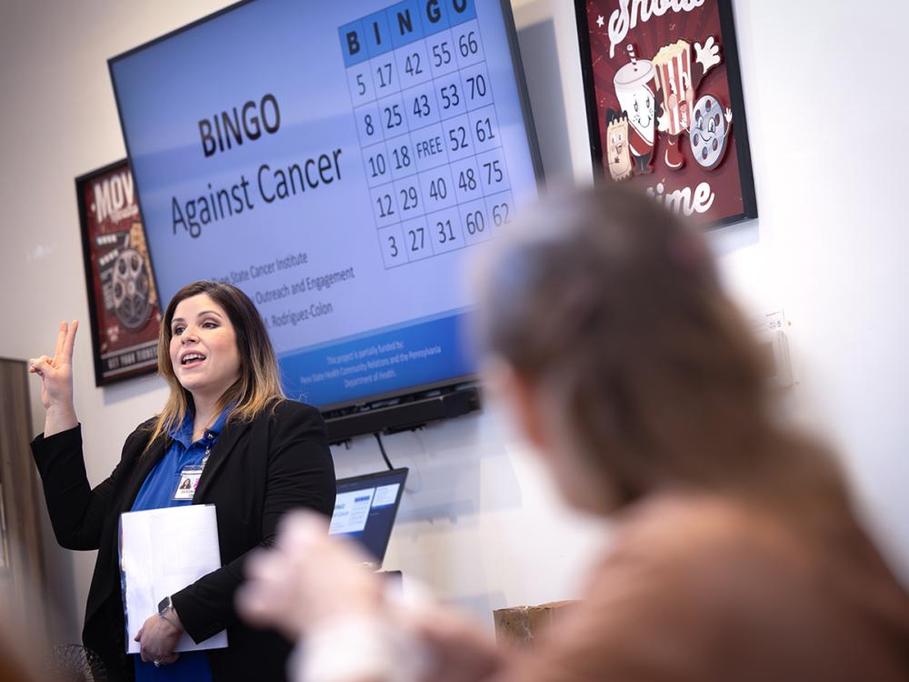 A woman stands in front of a screen with the words “BINGO Against Cancer” on it and a bingo card. She puts two fingers in the air. A woman, out of focus on the right, watches her.