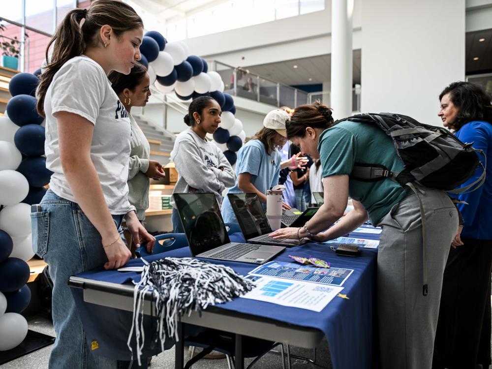 A person with a backpack leans over a table to type on a laptop while several students stand nearby at an indoor event, with informational materials on the table and blue-and-white balloon decorations in the background.