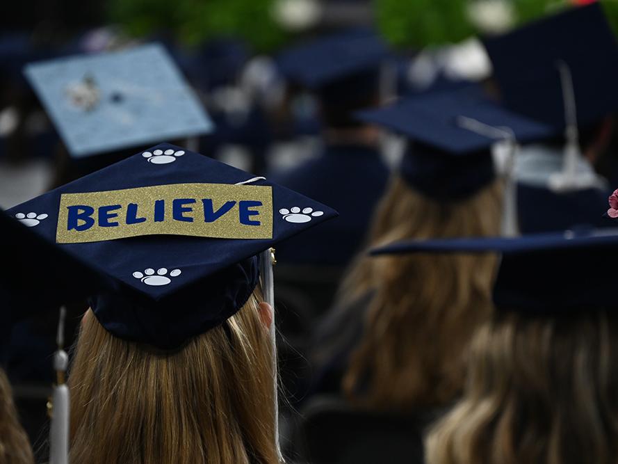 A graduate's mortar board displays the message "believe" during commencement