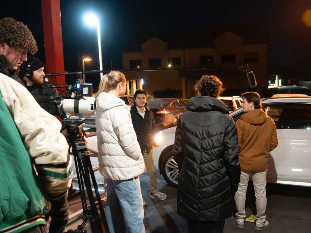 A group of people and a camera crew stand near a car.