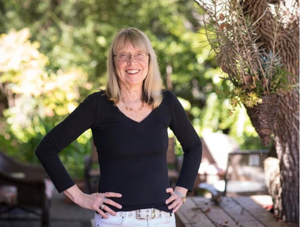 elderly person with blonde hair and glasses with trees in background