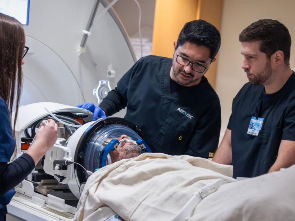Medical team preparing a patient for a focused ultrasound procedure, with clinicians adjusting equipment around the patient’s head inside a specialized imaging device.