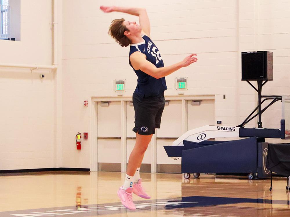 Caleb Fries jumps to serve a volleyball in a gymnasium, mid-air with arm extended, wearing a Penn State Altoona uniform.