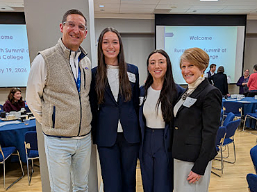 Four people, Mark Stephens, Kelsey Brown, Molly Graham and Lisa Davis, stand arm in arm smiling at the camera in a classroom with presentation slides in the background.