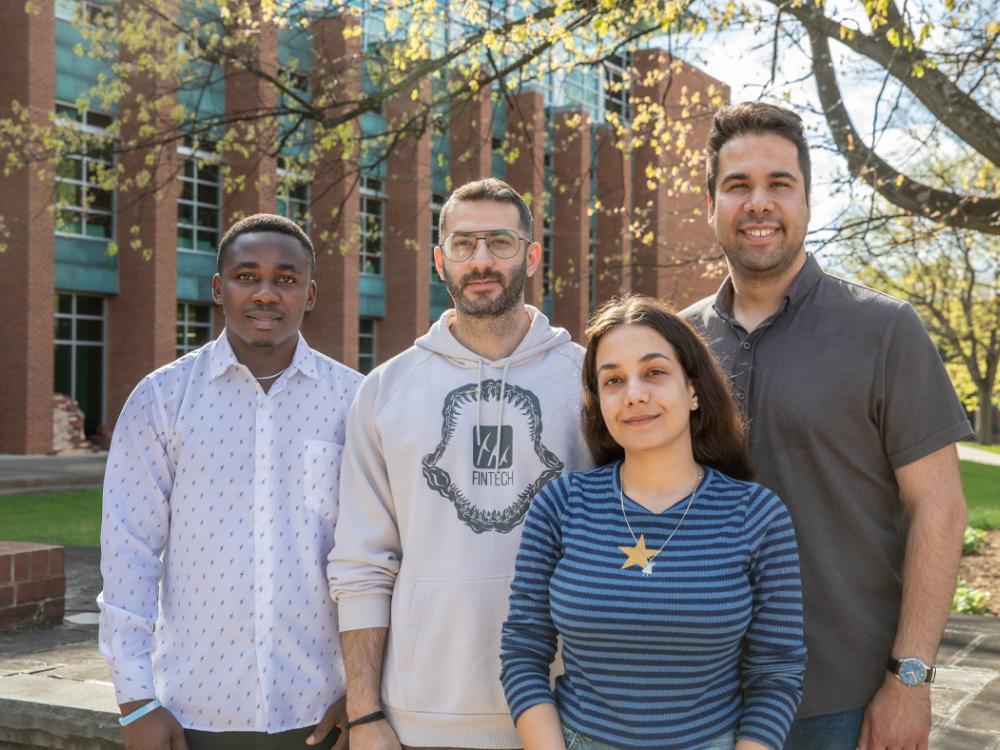 Four people stand together outdoors in front of a brick and glass campus building, with trees overhead. They are casually dressed, facing the camera, and positioned closely in a small group on a sunny day.