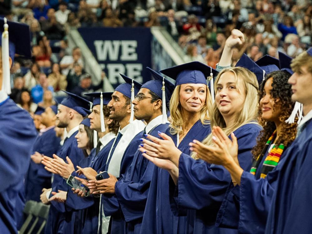 college students in blue caps and gowns applaud at commencement ceremony