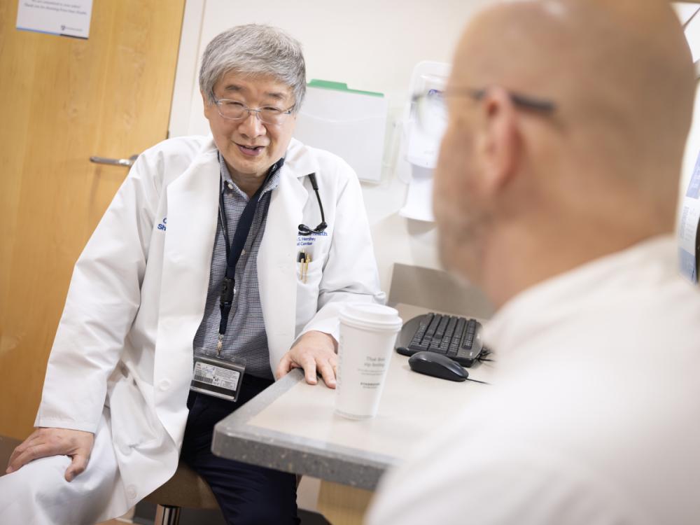 A physician wearing a white medical coat and a nametag is seated sideways in a clinic room, speaking with a patient who is in the foreground, slightly out of focus and looking away from the camera.