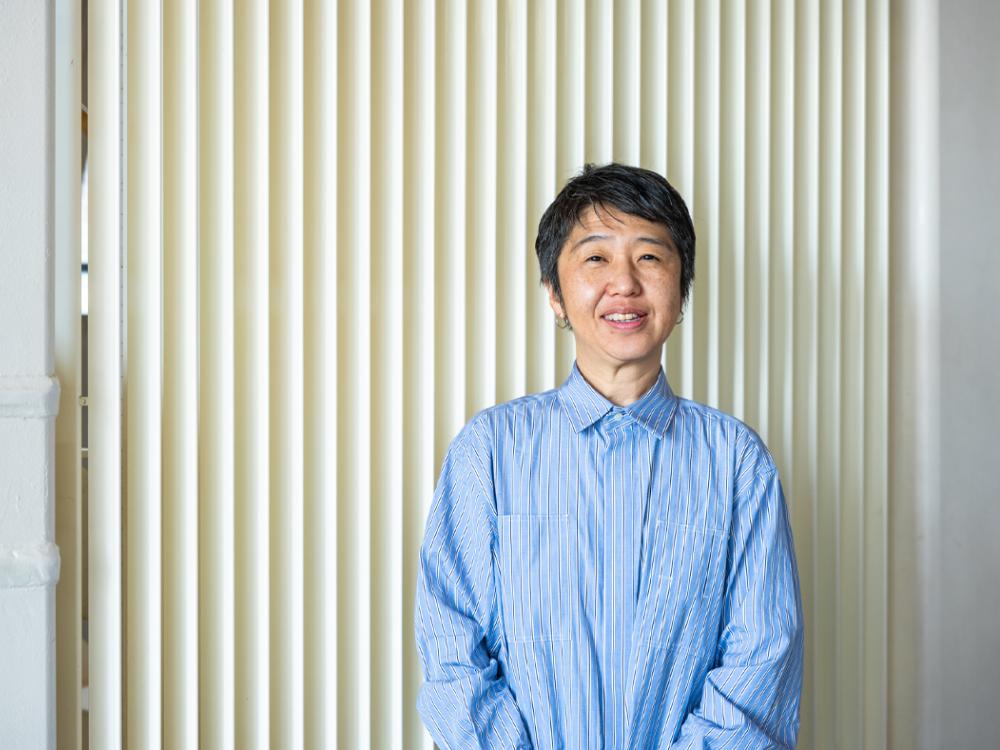 A person in an oversized blue shirt with ivory blinds as a backdrop.