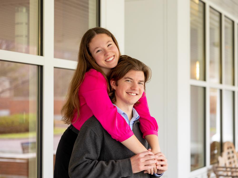 Sabrina, wearing a hot pink blouse, and Morgan, wearing a dark gray quarter-zip and blue button-up, pose together outside a wall of windows. 