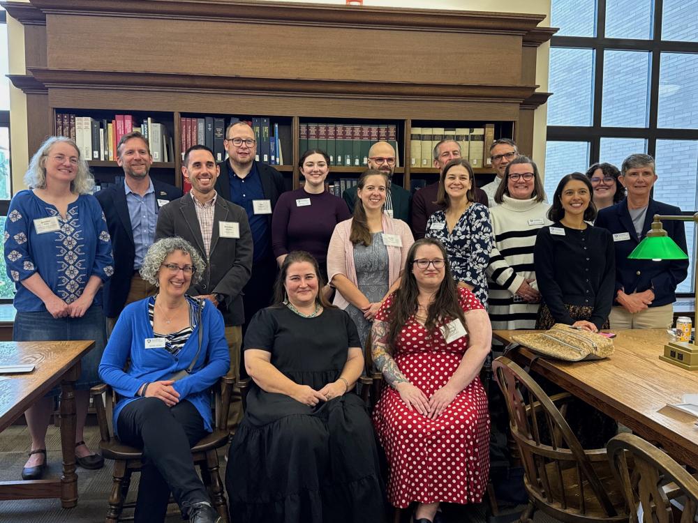 group photo of 17 people 14 standing 3 sitting behind library bookshelves and near large study tables