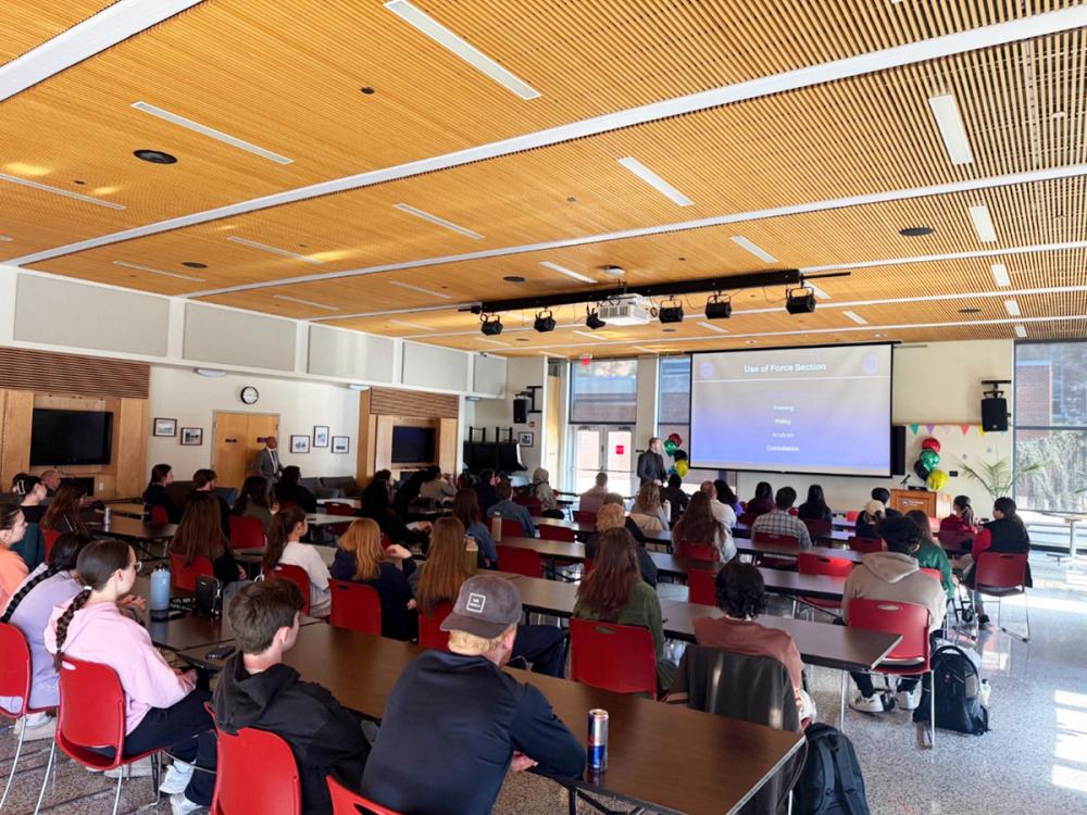 Students sit at tables in a classroom watching a presenter speak at the front, with a projected slide displayed on a large screen.
