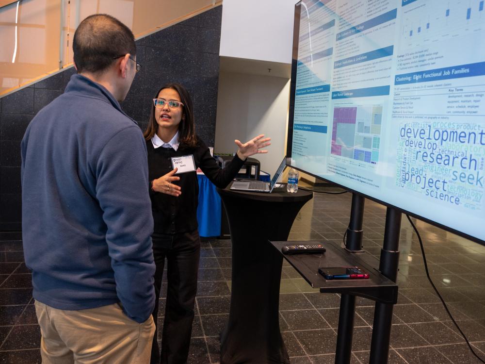 A student talks with a guest about her research poster, gesturing to it on a screen.