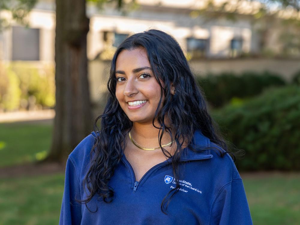 Mayuri Purani smiles and wears a blue Penn State College of the Liberal Arts peer adviser quarter-zip sweatshirt while standing outdoors on campus. 