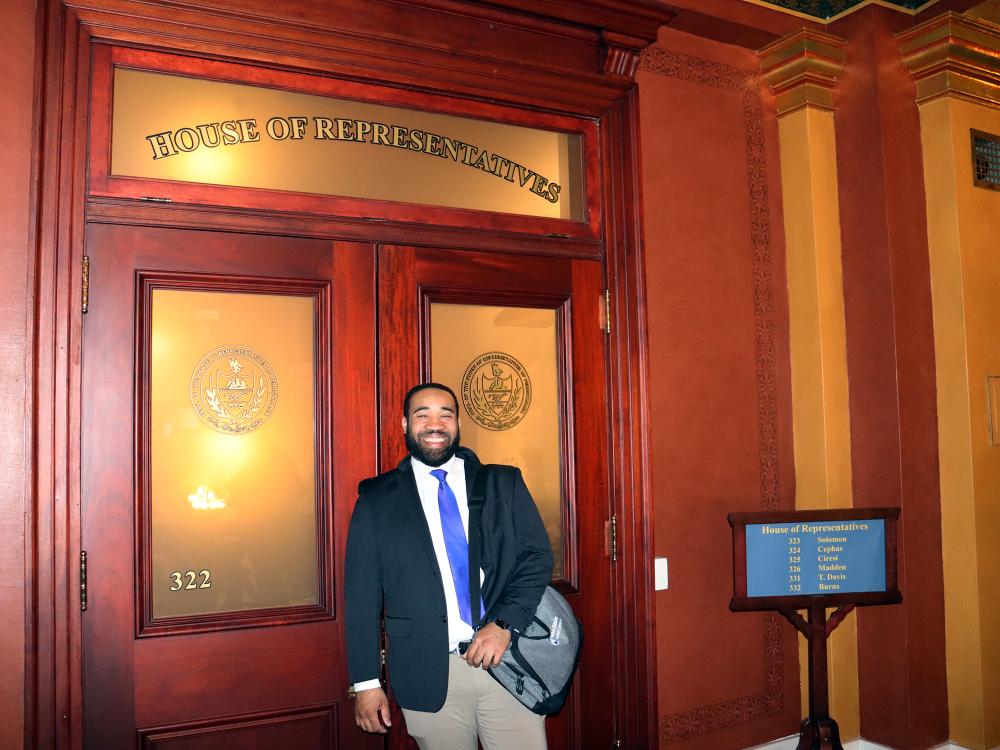 A man wearing a suit stands in front of a door that reads "House of Representatives."