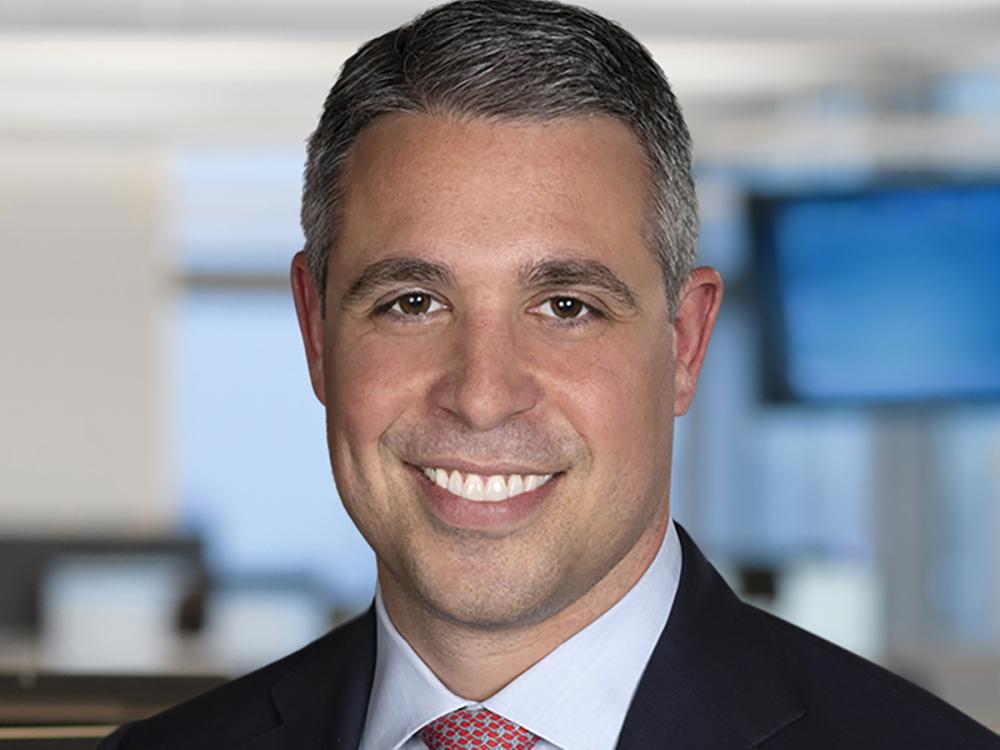 A head shot of a smiling Ryan Newman wearing a black suit coat and red patterned tie under a white shirt.