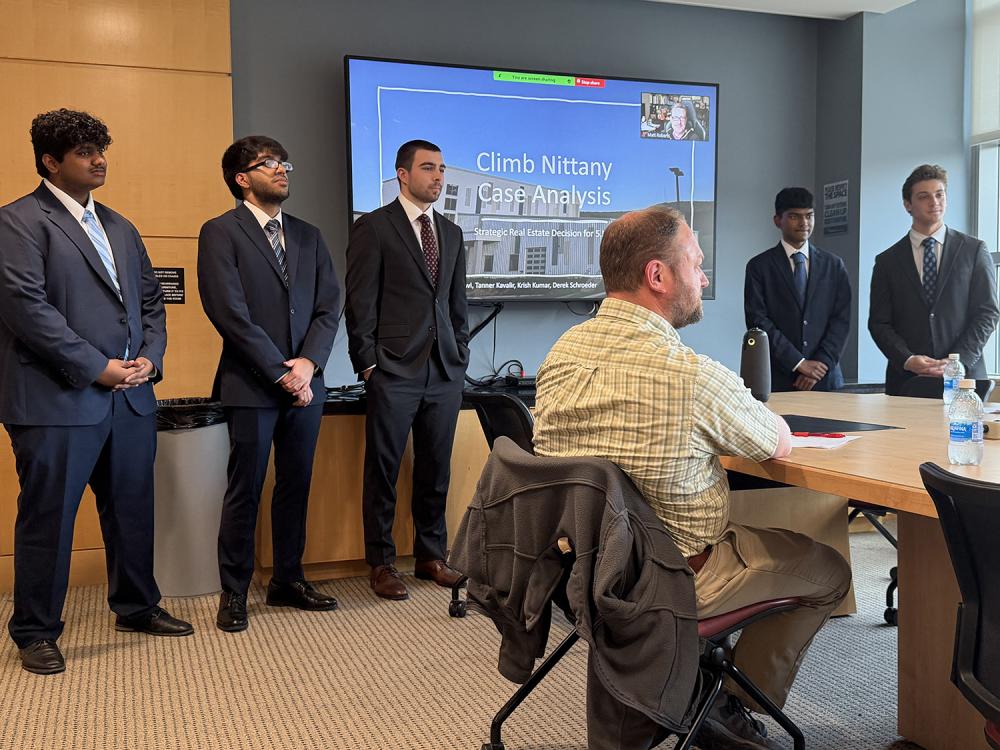 A team of five male students stands in front of a monitor in a conference room with judges seated at a table