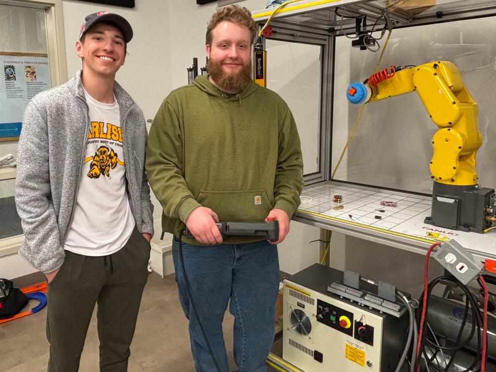 Two students stand next to a large yellow robotic arm on a table