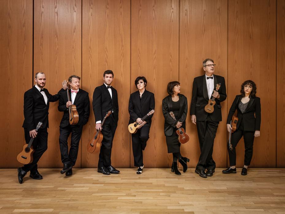 Men and women wearing tuxedos and holding a ukulele stand against a wood-paneled wall.