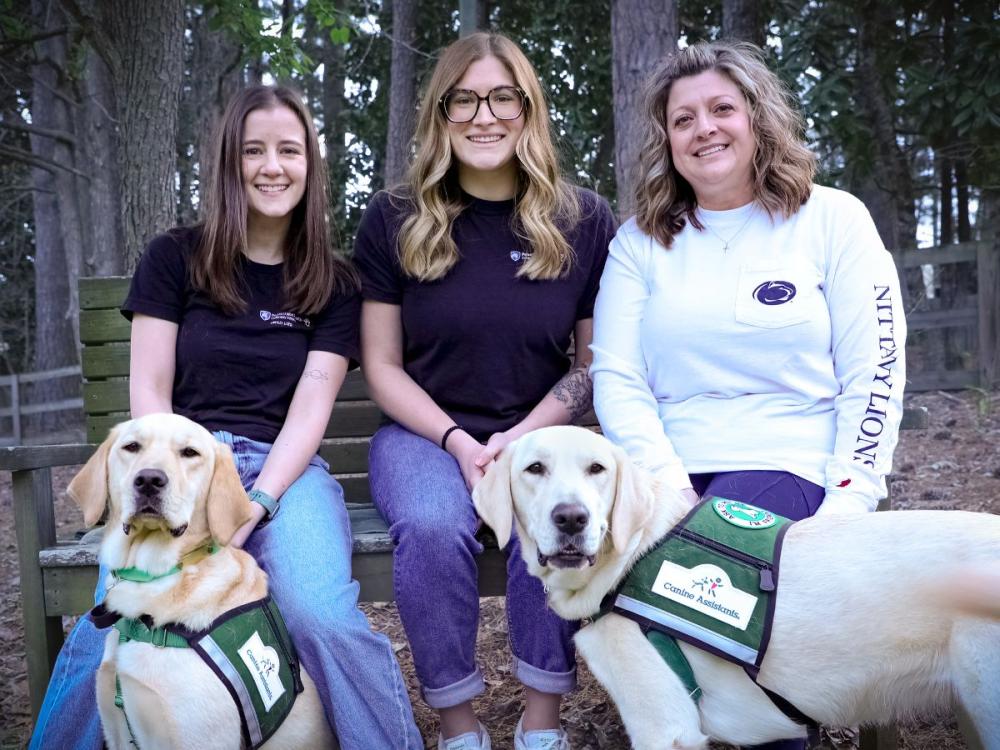 Three women sit on a wooden bench outdoors in a wooded area, smiling at the camera, each accompanied by a Golden Labrador retriever wearing a green “Canine Assistants” service vest.