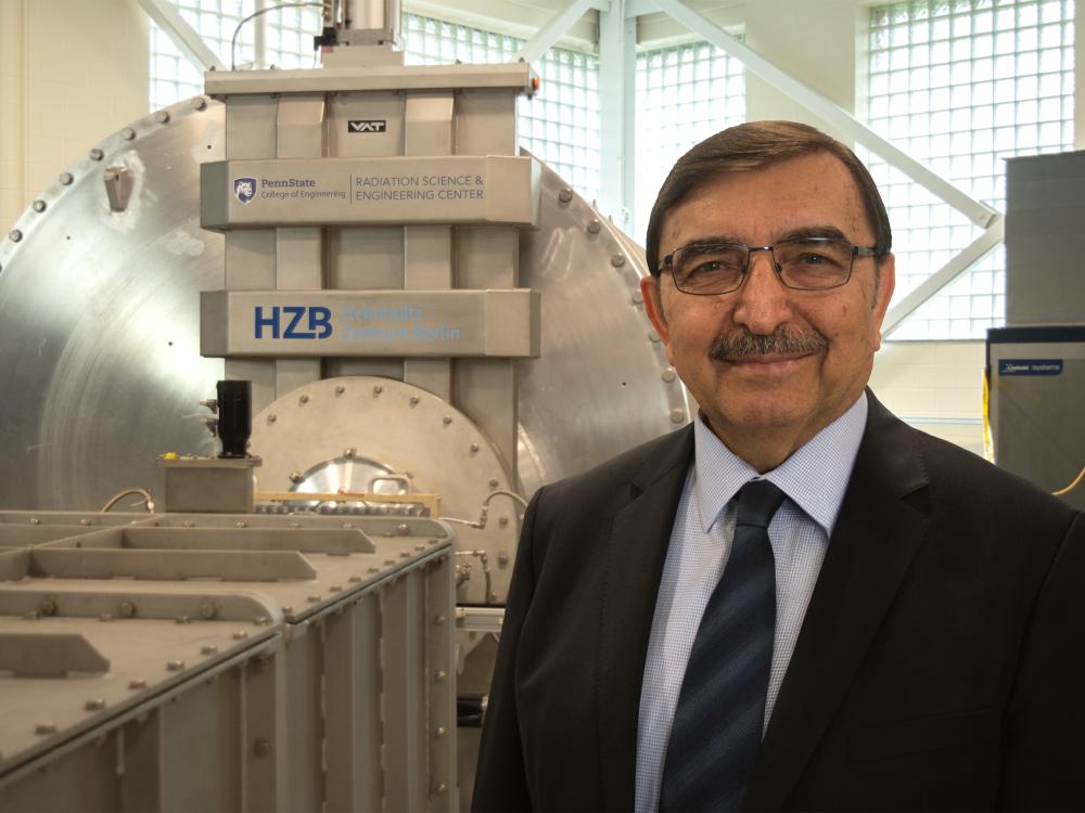A person wearing a suit jacket, tie, and glasses smiles at the camera in front of metal machinery. 