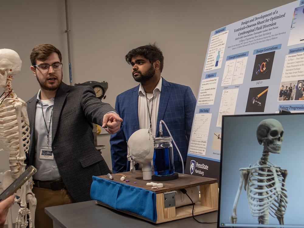 Two students, standing near a poster and a skeleton, explain their capstone project to guests